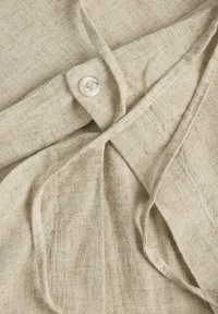 Bombažna srajca v svetlo bež barvi z gumbnim zapiranjem in veznim detajlom. Tkanina se zdi teksturirana z naravno, slubo končano površino.