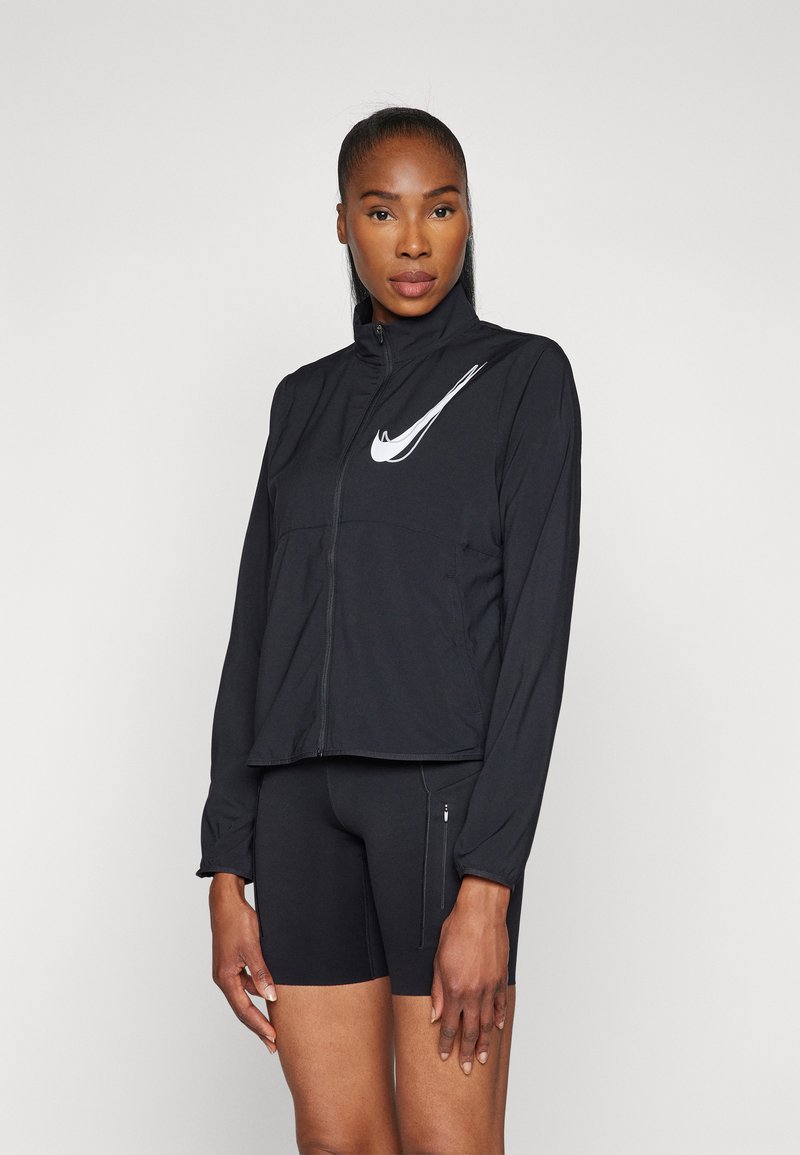 Sort Nike zip-up jakke med høj krave, der har et hvidt swoosh-logo på venstre side. Styliseret med sorte tætsiddende shorts for et sporty look.