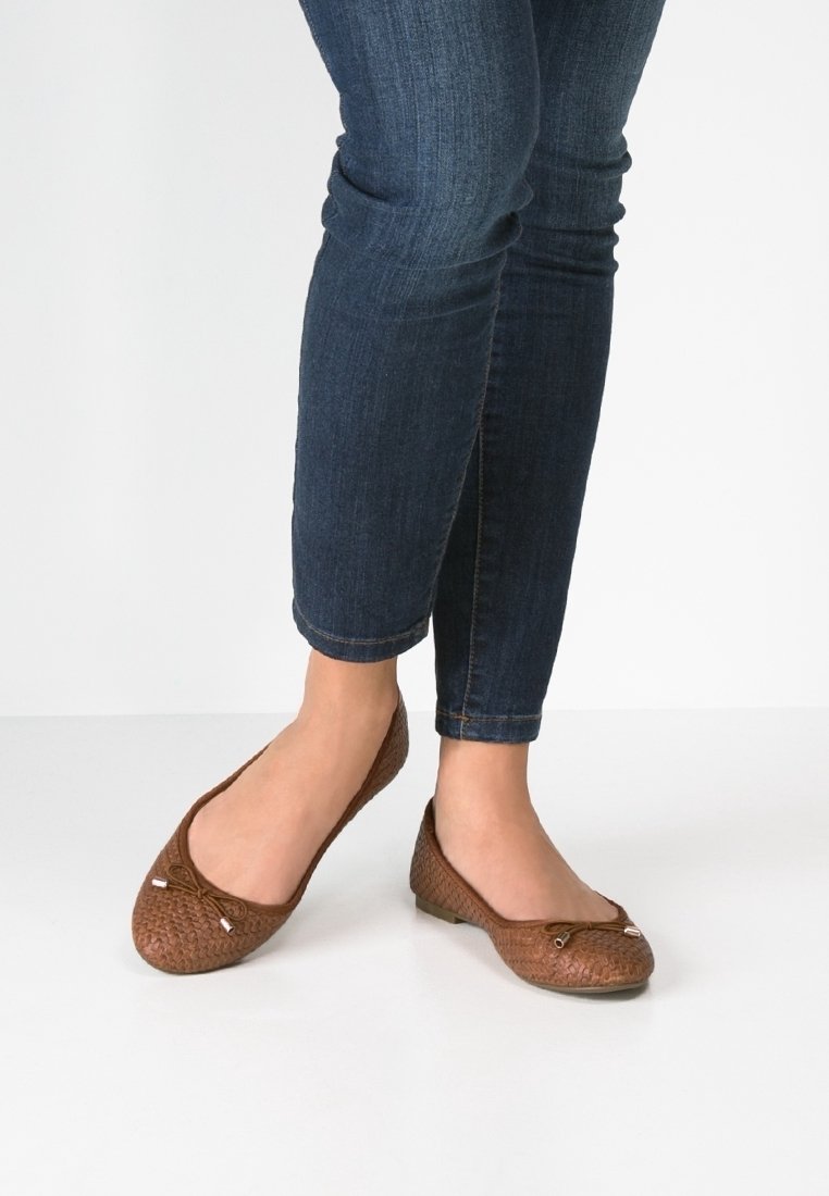 Pieds chaussés de ballerines brunes tissées avec de petits nœuds, associées à un jean skinny bleu foncé retroussé, sur un sol blanc.