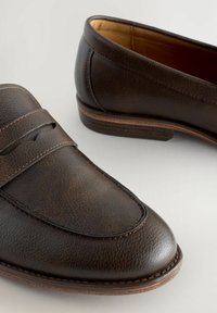 Mocasines de cuero marrón oscuro con una superficie texturizada, detalles de costura y un interior suave. Tacón ligeramente elevado para mayor soporte.