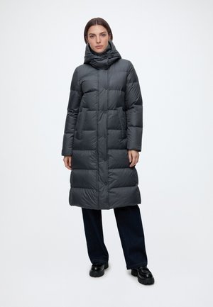 Mujer de pie vistiendo un abrigo de invierno acolchado largo de color gris oscuro con cuello alto, pantalones negros y zapatos negros con suela gruesa sobre fondo liso.