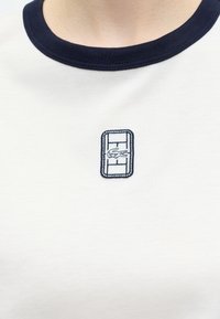 T-shirt blanc col rond avec col bleu marine et petit écusson rectangulaire avec un logo crocodile brodé au centre de la poitrine.