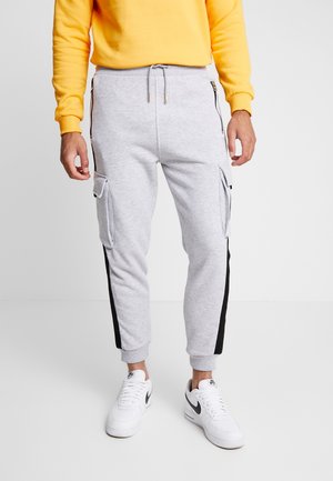 Graue Sweatpants mit schwarzen seitlichen Streifen, elastischem Bund und zwei seitlichen Cargo-Taschen. Getragen mit einem gelben Sweatshirt und weißen Turnschuhen.