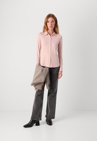 Lyserød skjorte med krave, parret med højtaljede, mørkegrå, brede bukser. Model holder en ternet blazer. Sorte ankelstøvler.