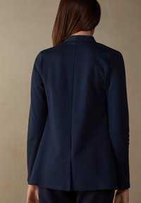 Námořnický modrý blazer, hladká tkanina, přiléhavý střih, dlouhé rukávy, detal na zadní straně, minimalistický design bez viditelného kovového příslušenství.