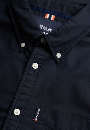 Granatowa koszula na guziki z białymi guzikami, zapinanym kołnierzykiem, kieszenią na piersi oraz małą metką z logo Superdry nad kieszenią.