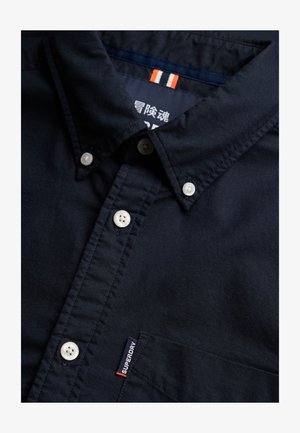 Granatowa koszula na guziki z białymi guzikami, zapinanym kołnierzykiem, kieszenią na piersi oraz małą metką z logo Superdry nad kieszenią.