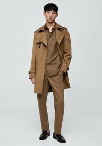 Cappotto trench marrone sopra una camicia a bottoni e pantaloni coordinati, con una vestibilità sartoriale e una texture della stoffa liscia. Scarpe nere completano il look.