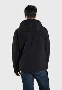 Giacca softshell nera con cappuccio, dalla trama liscia e dal fit classico. Abbinata a jeans blu, evidenziando il suo design versatile.