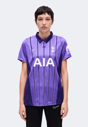 Άτομο που φοράει μωβ φανέλα ποδοσφαίρου της Tottenham Hotspur με λευκό λογότυπο "AIA" και μαύρο παντελόνι Nike, στέκεται σε απλό φωτεινό φόντο.