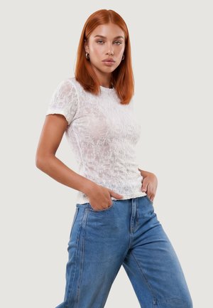 Mujer joven con cabello rojo que lleva una blusa blanca de encaje de manga corta y vaqueros azules con las manos en los bolsillos sobre un fondo liso.