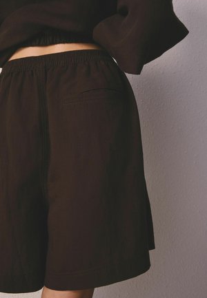 Taille und obere Beine einer Person, die lockere schwarze Shorts mit elastischem Bund und Taschen-Detail trägt, vor einer strukturierten hellgrauen Wand.