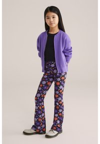 Cardigan violet avec fermeture éclair, chemise noire et leggings noirs évasés ornés de motifs colorés de marguerites. Baskets blanches avec semelles noires.