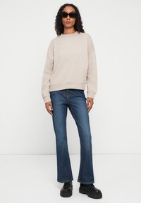 Mujer con cabello rizado, llevando gafas de sol negras, sudadera beige de manga larga, jeans de corte bootcut azules y zapatos de plataforma negros, de pie.