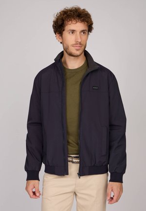 Homme aux cheveux bouclés portant une veste noire zippée par-dessus un pull vert olive et un pantalon beige, debout devant un fond uni.