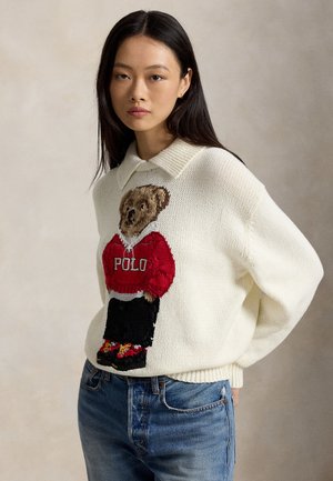 Femme aux longs cheveux foncés portant un pull blanc avec un ours en sweat-shirt Polo rouge et un jean bleu, debout devant un fond neutre.