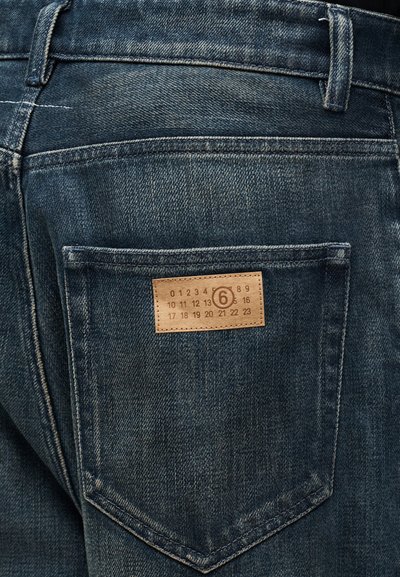 Poche arrière d'un jean en denim bleu foncé avec une petite étiquette en cuir marron affichant des chiffres disposés en rangées et un chiffre 6 encerclé.