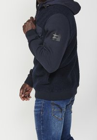 Mörkblå zip-up sweatshirt med en texturerad fleecekropp och släta ärmar, med en bröstficka och logopatch på ärmen.