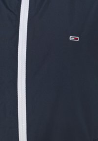 Navy blue jacket with white zipper and small rectangular logo featuring white, red, and navy blue stripes on the chest.