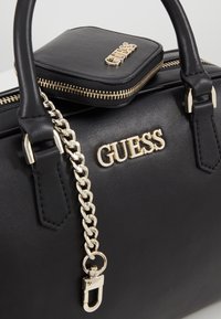 Sac à main en cuir noir avec détail chaîne dorée et logo GUESS en lettres dorées sur le devant, ainsi qu'une petite pochette assortie fixée par une fermeture éclair dorée.