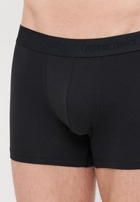Schwarze Trunks aus Bio-Baumwolle mit einem weichen Bund, der den Text "Organic Basics" trägt. Das Design ist eng geschnitten und hat eine glatte Textur.