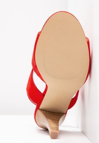 Sandale à talon haut en daim rouge avec un dos ouvert et des lanières latérales, dotée d'une semelle beige texturée et d'un talon en bois effilé.