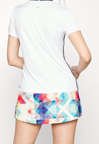 Bílé sportovní tričko s krátkým rukávem, doplněné pestrou sukní s geometrickým vzorem v odstínech modré, růžové a oranžové.
