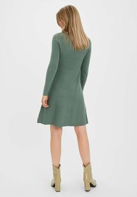 Femme aux cheveux blonds portant une robe verte à manches longues et hauteur genoux, avec des bottines beiges à la cheville, se tenant de dos par rapport à l'appareil photo.