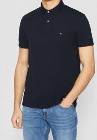 Man som bär en figursydd marinblå polo med en liten logotyp på bröstet och ljusblå jeans, en hand i fickan.