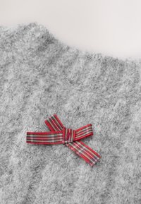 Pull en maille grise à la texture douce, avec un nœud à carreaux rouge et vert en accent sur le côté gauche.