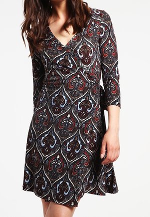 Kobieta ubrana w sukienkę sięgającą kolan z dekoltem w kształcie litery V, o rękawach długości do łokcia i wielokolorowym wzorze paisley w kolorach czerwonym, brązowym, niebieskim i czarnym.