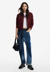 Burgunderfarbene Leder-Bomberjacke über einem weißen, cropped T-Shirt, mit blauen Patchwork-Weitbeinjeans und schwarzen Sneakern, während eine schwarze Handtasche gehalten wird.