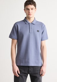 Polo shirt bleu gris avec un col, deux boutons et un petit patch logo noir sur la poitrine. Manches courtes, en coton.