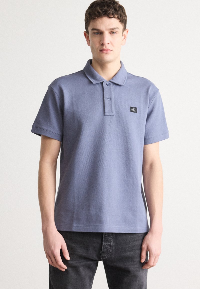Polo shirt bleu gris avec un col, deux boutons et un petit patch logo noir sur la poitrine. Manches courtes, en coton.