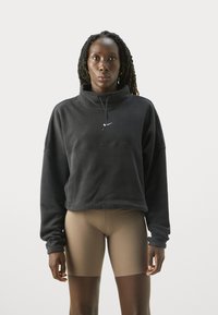 Kvinde med flettede hår, iført en sort Nike pullover og beige cykelshorts, stående mod en ensfarvet hvid baggrund.
