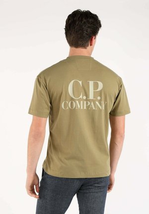 Uomo che indossa una T-shirt verde oliva a maniche corte con grande scritta bianca "C.P. Company" sulla schiena, abbinata a jeans scuri.