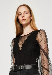 Body nero con maniche a pois trasparenti, dettagli in pizzo floreale e scollatura a V, abbinato a una cintura larga nera e jeans scuri.