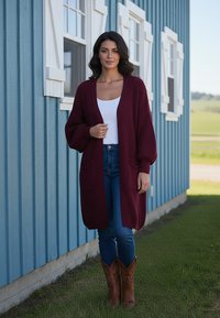 Burgundowa dziergana kardigan z luźnymi rękawami i bocznymi kieszeniami, w zestawieniu z niebieskimi dżinsami i brązowymi kowbojkami, na tle niebieskiej ściany.