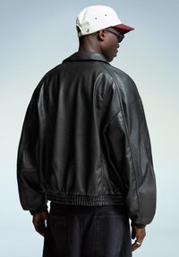 Giubbotto bomber in pelle nera con orlo e polsini a coste, caratterizzato da una finitura testurizzata. In testa è indossato un cappellino bianco e bordeaux.