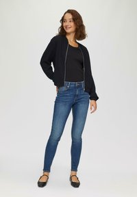 Schwarze Bomberjacke mit geripptem Muster, kombiniert mit engen blauen Jeans und schwarzen Ballerinas. Das Outfit ist lässig und schmal geschnitten.