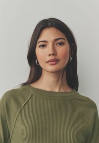 Pull en maille côtelée vert olive avec un col rond, doté de manches raglan et d'un tissu texturé. Le mannequin a des cheveux foncés de longueur moyenne.