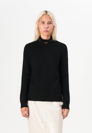 Black ribbed knit sweater with a high neck and a round gold hardware accent. Worn over a light textured skirt.