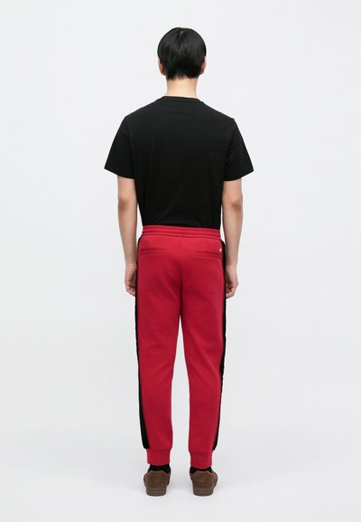 Punaiset collegehousut mustilla sivuraidoilla, joustavalla vyötäröllä ja kahdella takataskulla. Malli pukeutuneena mustaan paitaan ja ruskeisiin kenkiin.