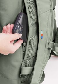 Vihreä kangaskansio, jossa on verkkotasku; käsi asettaa puhelimen sivutaskuun. Varustettu säädettävillä olkahihnoilla ja pienillä väriyksityiskohdilla.