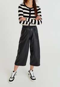 Schwarz-weiß gestreifter, gerippter Cardigan mit Knöpfen, kombiniert mit schwarzen, faux-ledernen, weit geschnittenen Hosen und Sneakers mit hohem Schaft.