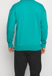 Sweat-shirt bleu sarcelle avec col rond, poignets et ourlet côtelés. Présente une texture lisse et des épaules tombantes. Associé à un pantalon noir.