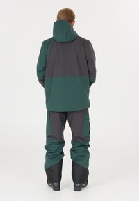 Veste et pantalon imperméables en vert foncé et gris, avec une capuche, des poches latérales et des poignets renforcés. Texture lisse, design pour extérieur.