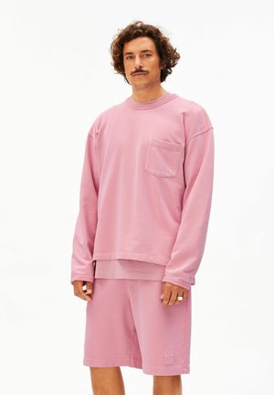 Mann mit lockigem Haar und Schnurrbart, der ein rosa Langarmhemd mit Brusttasche und passende rosa Shorts trägt und vor einem weißen Hintergrund steht.