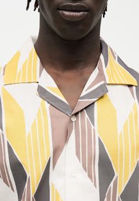 Άτομο με σκουρόχρωμο δέρμα που φορά κοντομάνικο πουκάμισο με κίτρινες, γκρι και μπεζ γεωμετρικές ρίγες σε λευκό φόντο, κουμπωμένο στο γιακά.