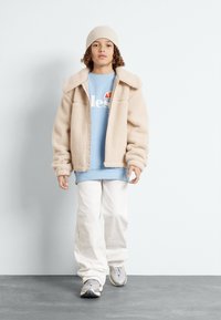 Chaqueta de pelo sintético beige con un cuello ancho, sudadera azul claro con logo, pantalones blancos holgados y zapatillas grises. Gorra de punto beige.